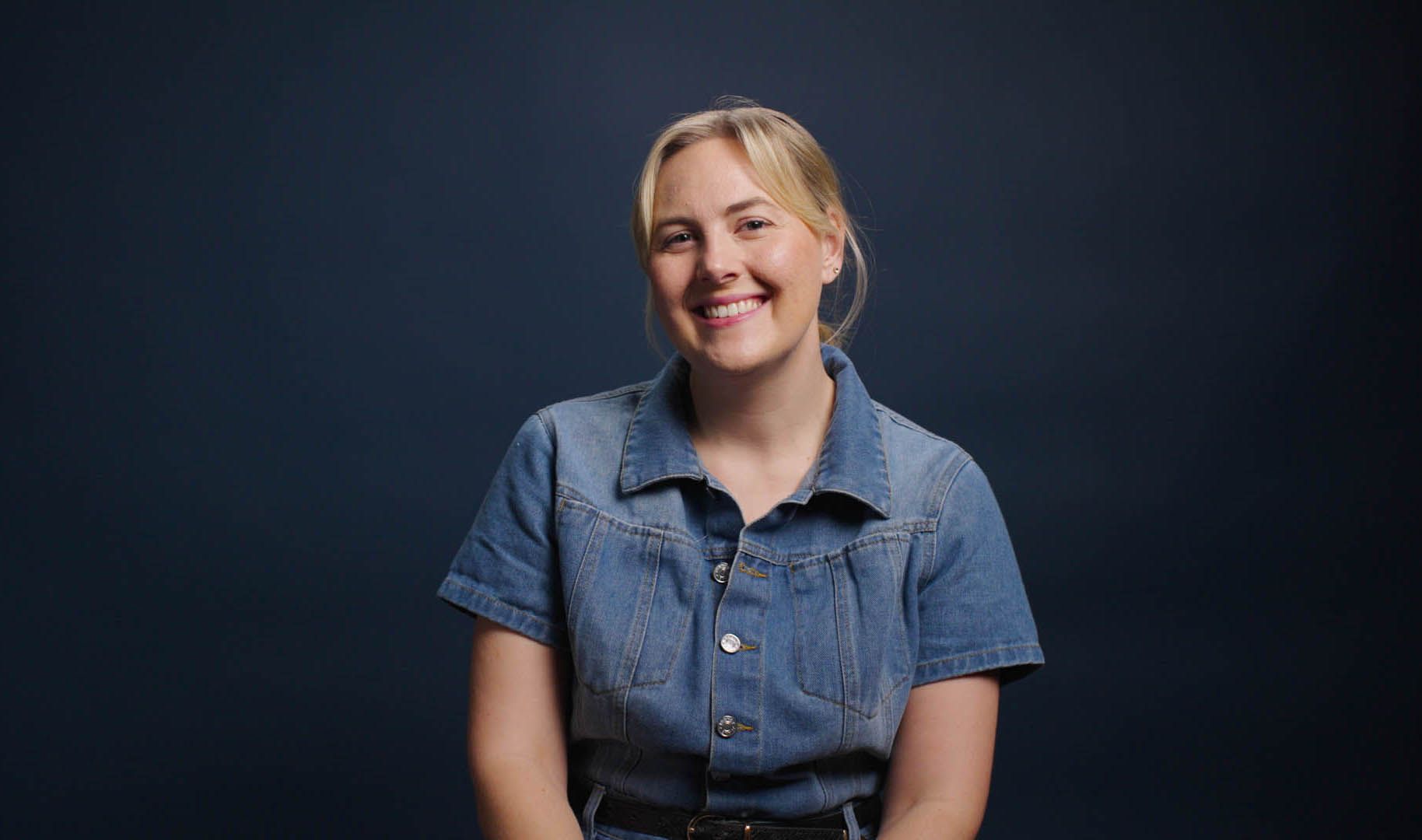 A woman in a denim dress is smiling and looking at the camera.