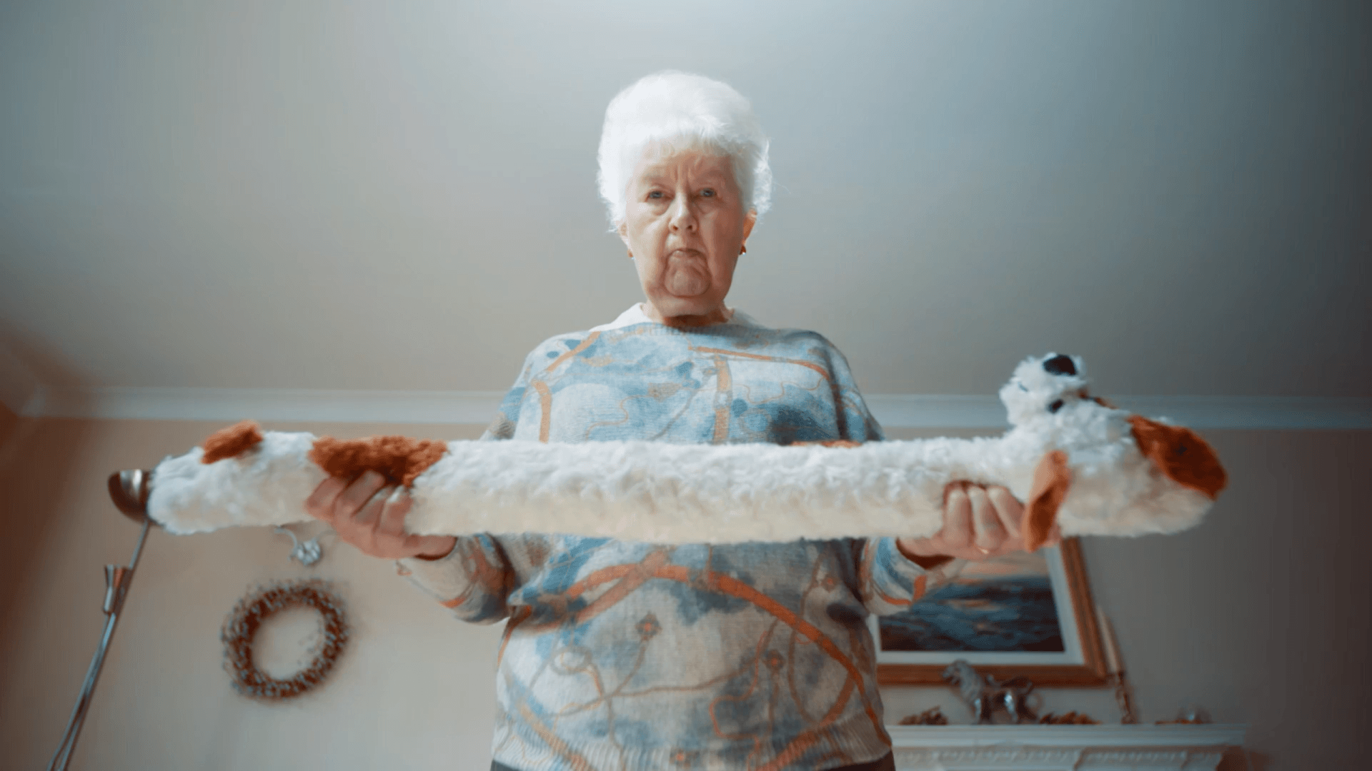 An elderly woman is holding a long stuffed animal in her hands.