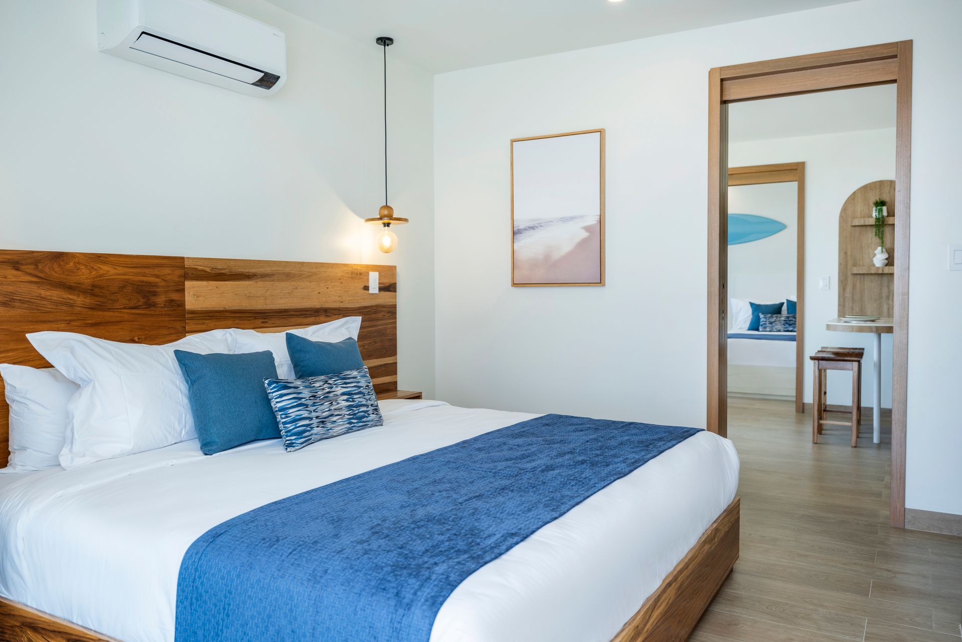 Modern bedroom with blue bedding, wood headboard, hanging lamp, and an open doorway to a bright adjoining room