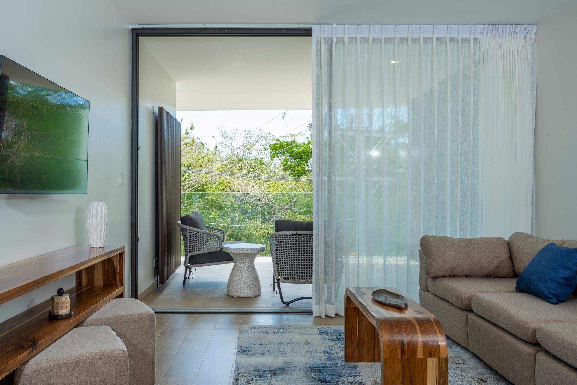 Modern living room with open balcony doors, sheer curtains, sofa, and greenery outside