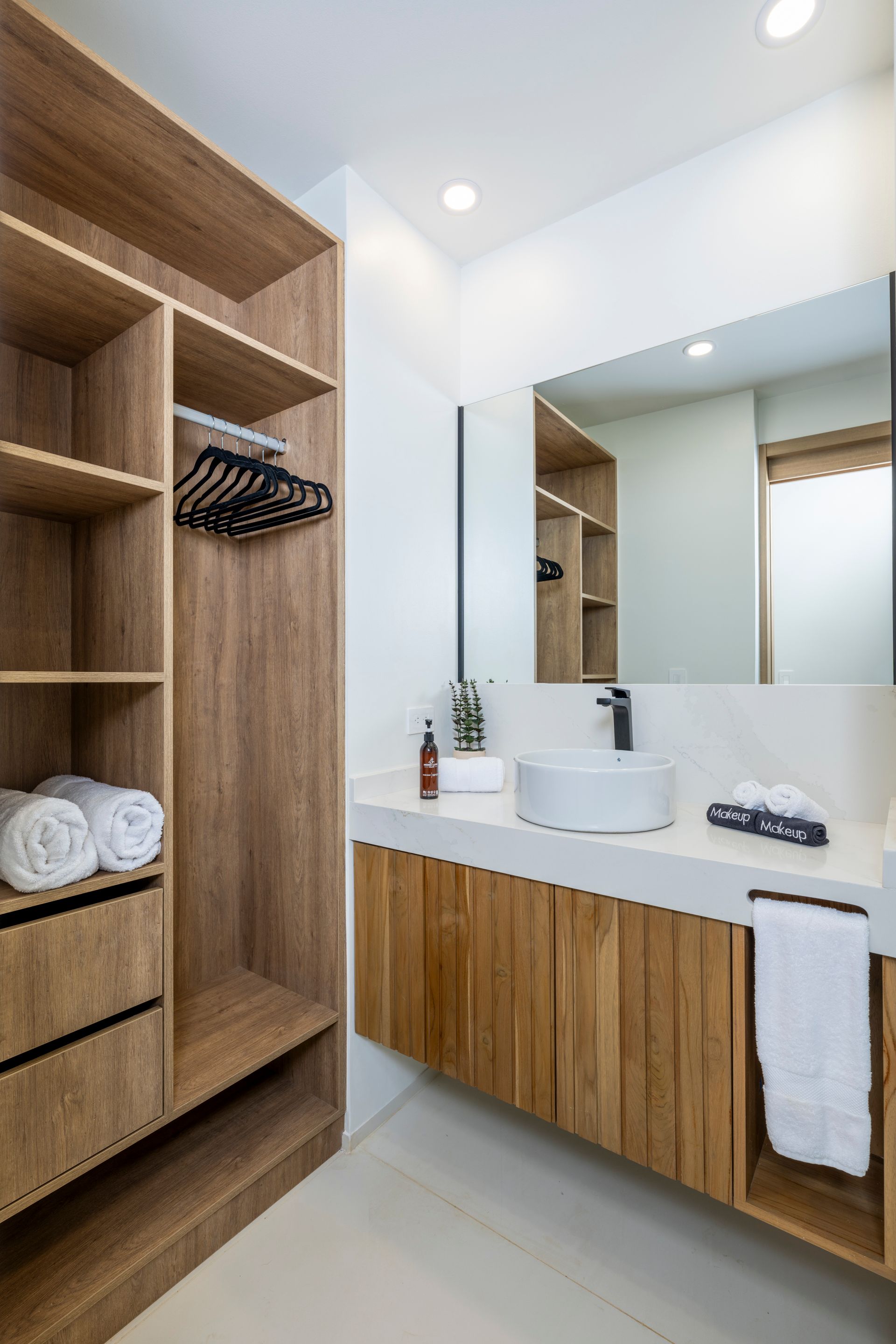 Modern bathroom with wood cabinetry, white sink, large mirror, and open shelving with towels and hangers