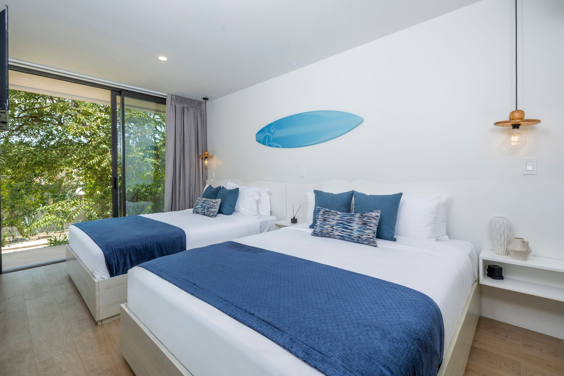 Bright modern bedroom with two white beds, blue bedding, wall art, and large windows overlooking greenery