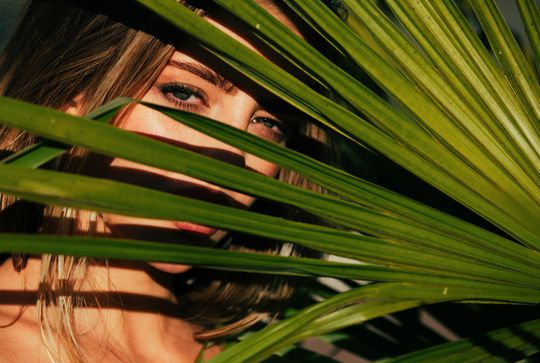 A close up of a woman 's face behind a palm tree leaf.