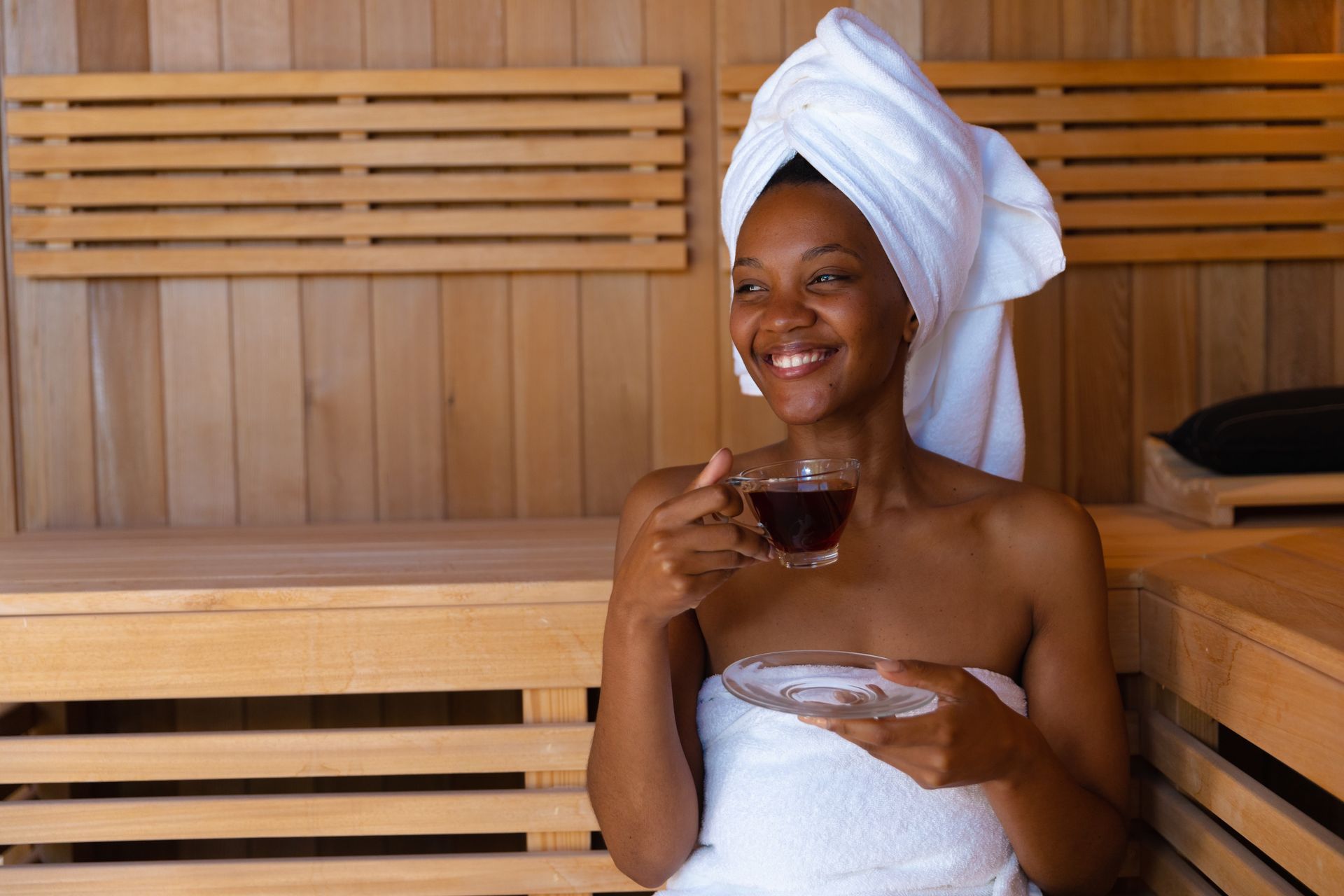 A woman wrapped in a towel is sitting in a sauna drinking a cup of tea.