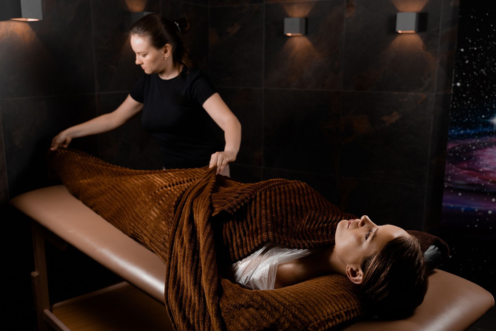 A woman is laying on a massage table wrapped in a brown blanket.