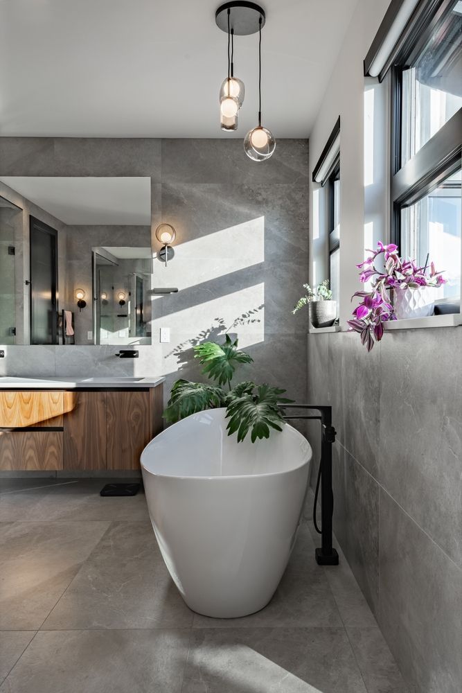 Modern bathroom with a white tub, plants, and grey tile. Natural light streams through the window.