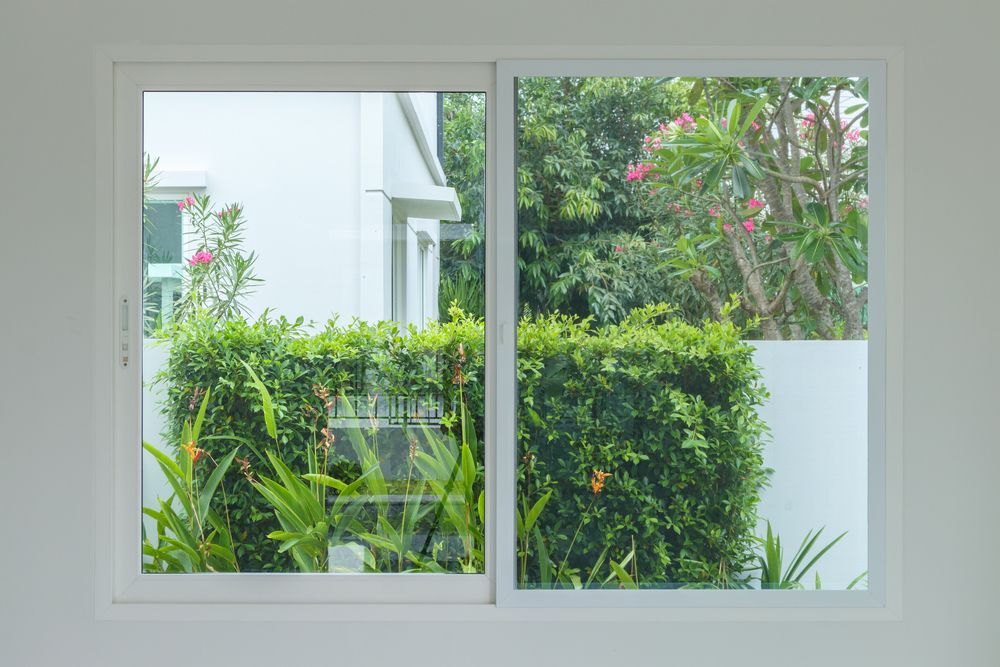 White-framed sliding window looking out at a lush garden and partially visible white house.