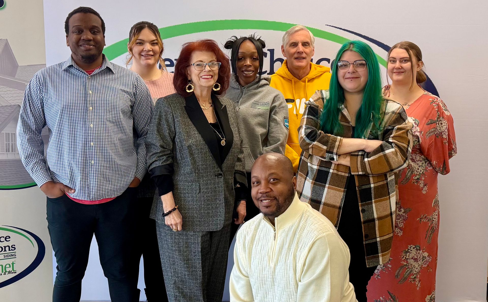 Group of people posing in front of a banner. Some are smiling. One person is kneeling. Indoor setting. At Perfect Choice Home Solutions.