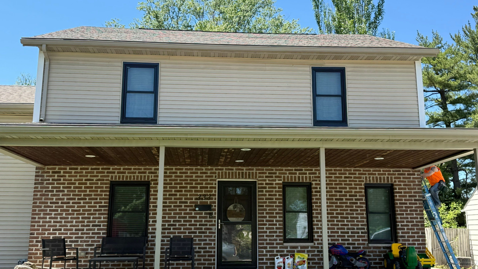 Two-story brick house with tan siding, black-trimmed windows, and a covered front porch under a blue sky
