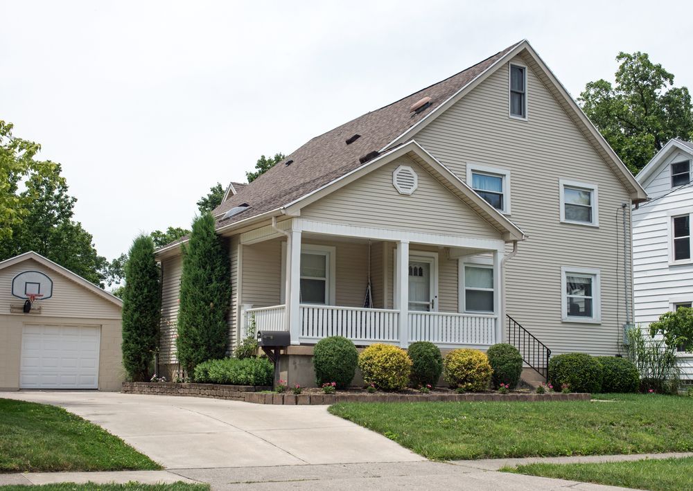 Two-story beige house with porch, attached garage with basketball hoop. Cloudy day, green lawn.
