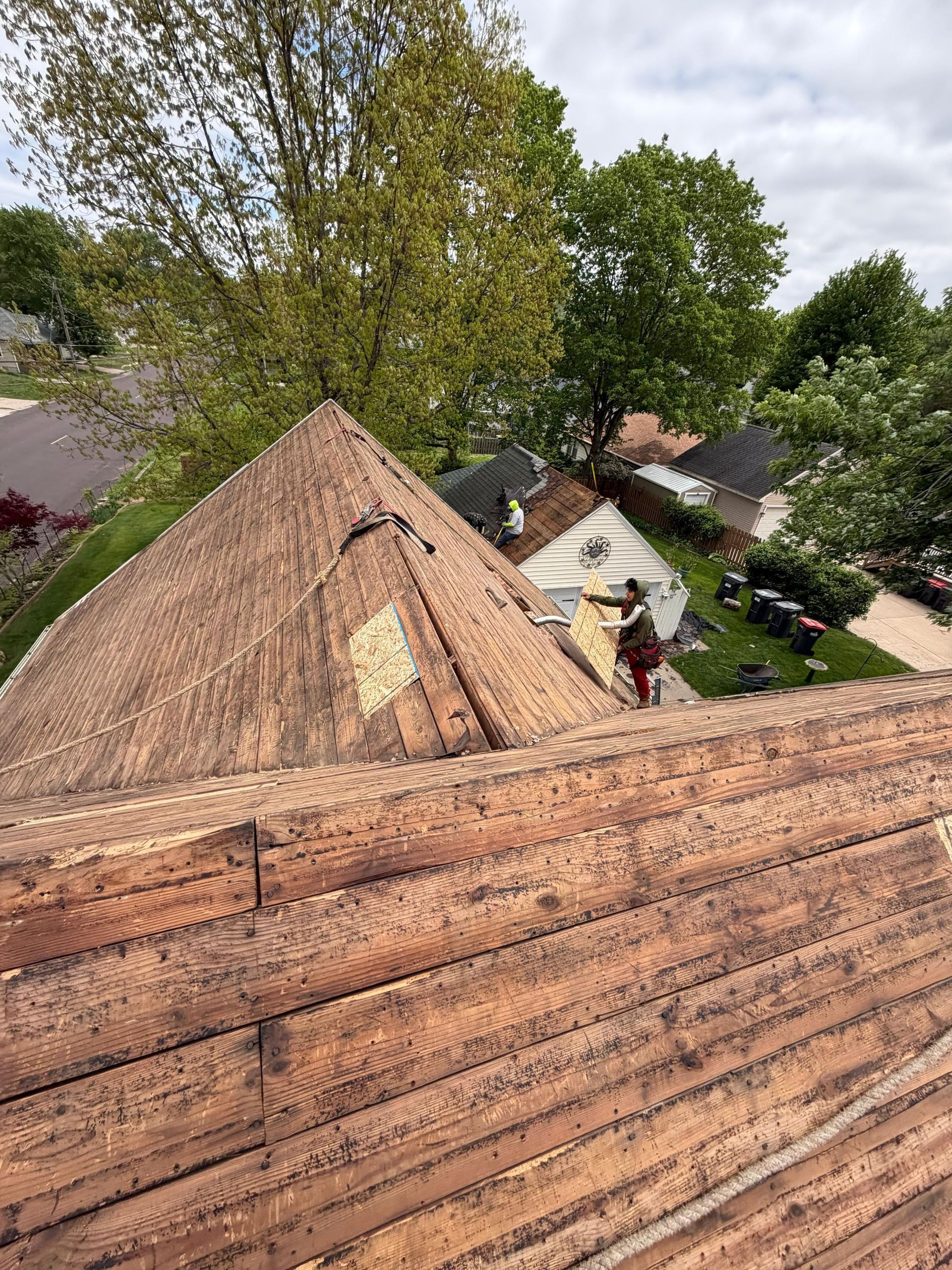 Roofer using a nail gun to install gray shingles on a rooftop. Worker wearing safety vest and gloves.