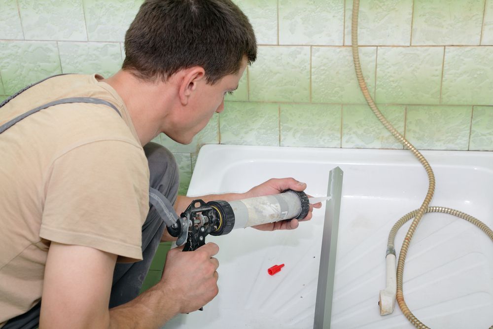 Person caulking a bathtub; a gray caulk gun is used. Green tiled wall in the background.