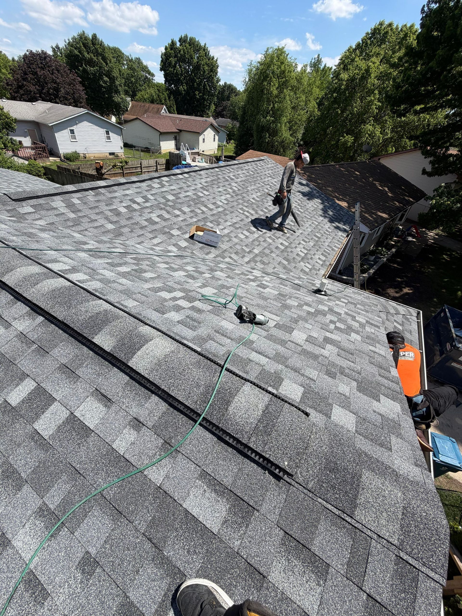 Roofers installing gray shingles on a steep residential roof under a blue sky