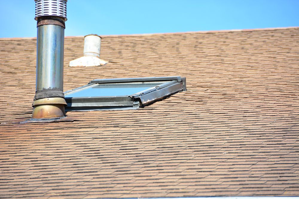Brown shingle roof with a metal chimney, skylight, and small vent. Sunny day with a blue sky.