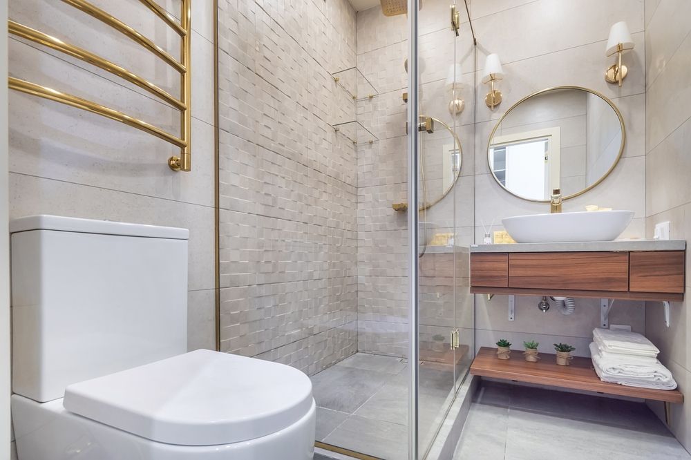 Modern bathroom with a white toilet, glass-enclosed shower, wood vanity, vessel sink, and gold fixtures on light tile.