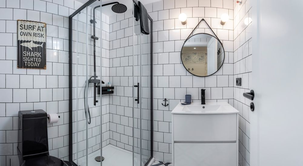 Modern bathroom with black accents, white tiled walls, glass shower, black toilet, and a round mirror.