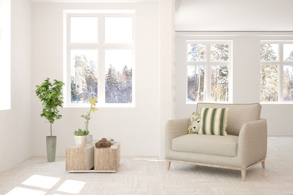 Bright, minimalist living room with beige armchair, small wooden table, and plants by a window overlooking a snowy forest.