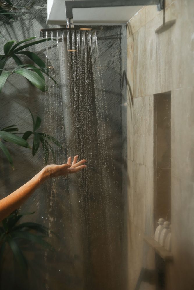 Hand under a large rain shower head in a tiled shower. Water streams down, with plants and a niche in the wall visible.