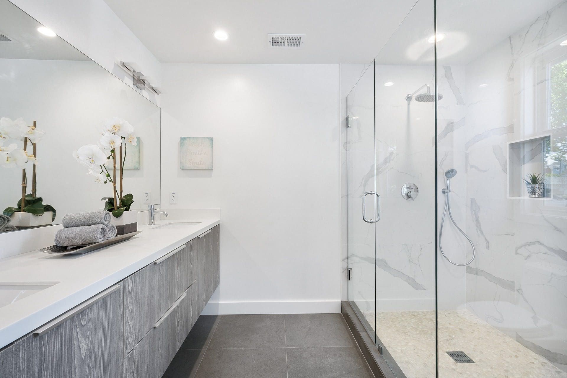 Modern bathroom with gray cabinets, white countertops, glass shower, and white marble tiles.