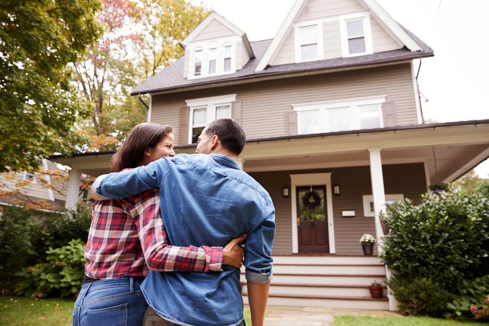 Couple with arms around each other looking at a brown house with a porch in a tree-lined yard.