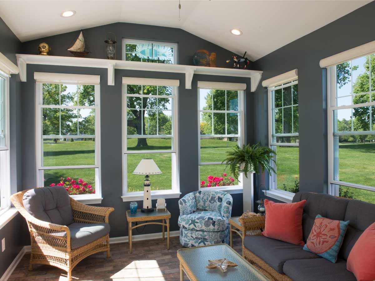 Sunroom with gray walls, large windows overlooking a lawn, and wicker furniture with colorful cushions.