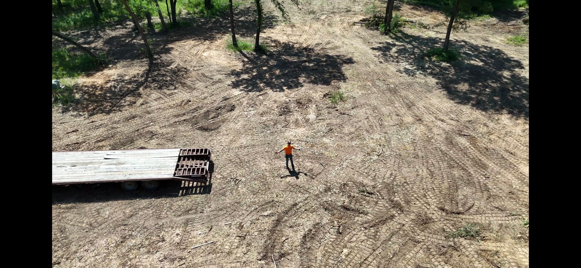 A worker in orange stands in a cleared area. A flatbed trailer is nearby, trees in the background.