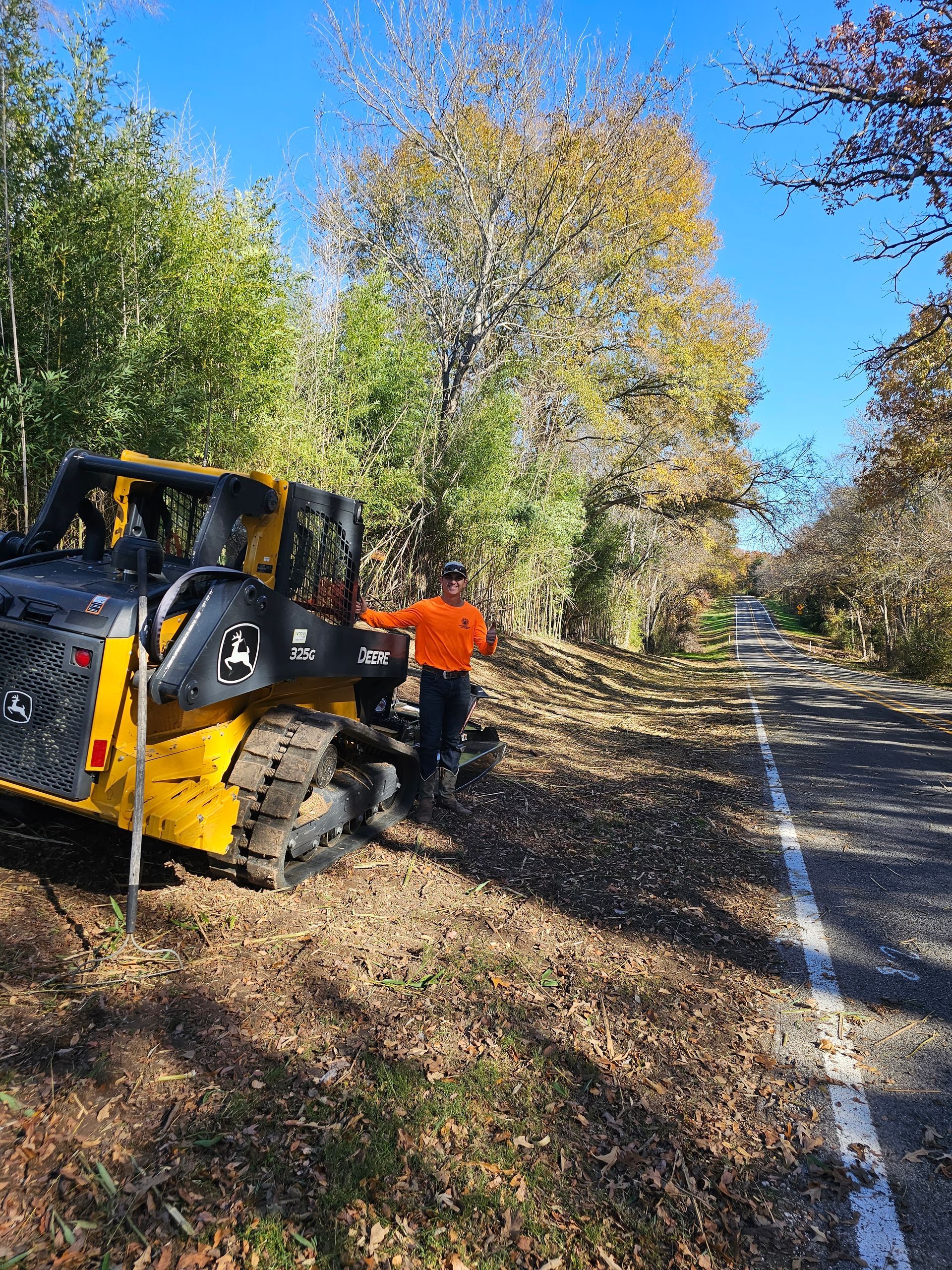 Man operating a yellow skid steer near a road, clearing brush with fall foliage.