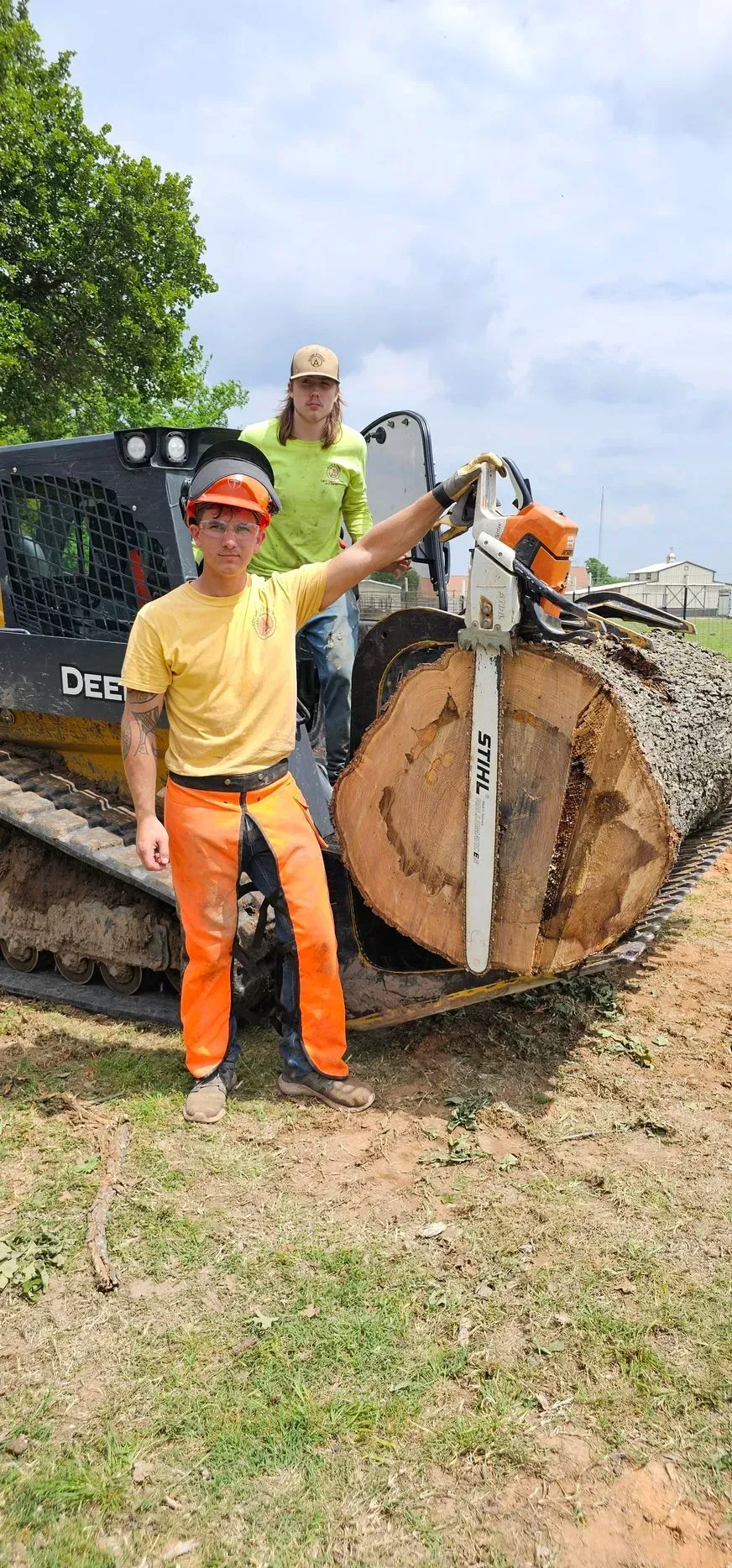 Two people with a chainsaw and a large log; one man points. Orange pants and a John Deere vehicle in the background.