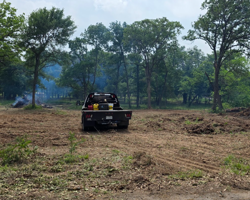 A pickup truck parked on cleared land, with trees in the background and smoke rising from a fire.