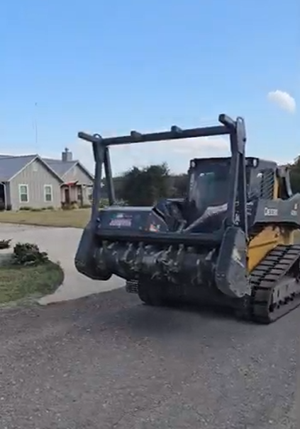 A John Deere skid steer with a brush cutter attachment on a driveway near a house.