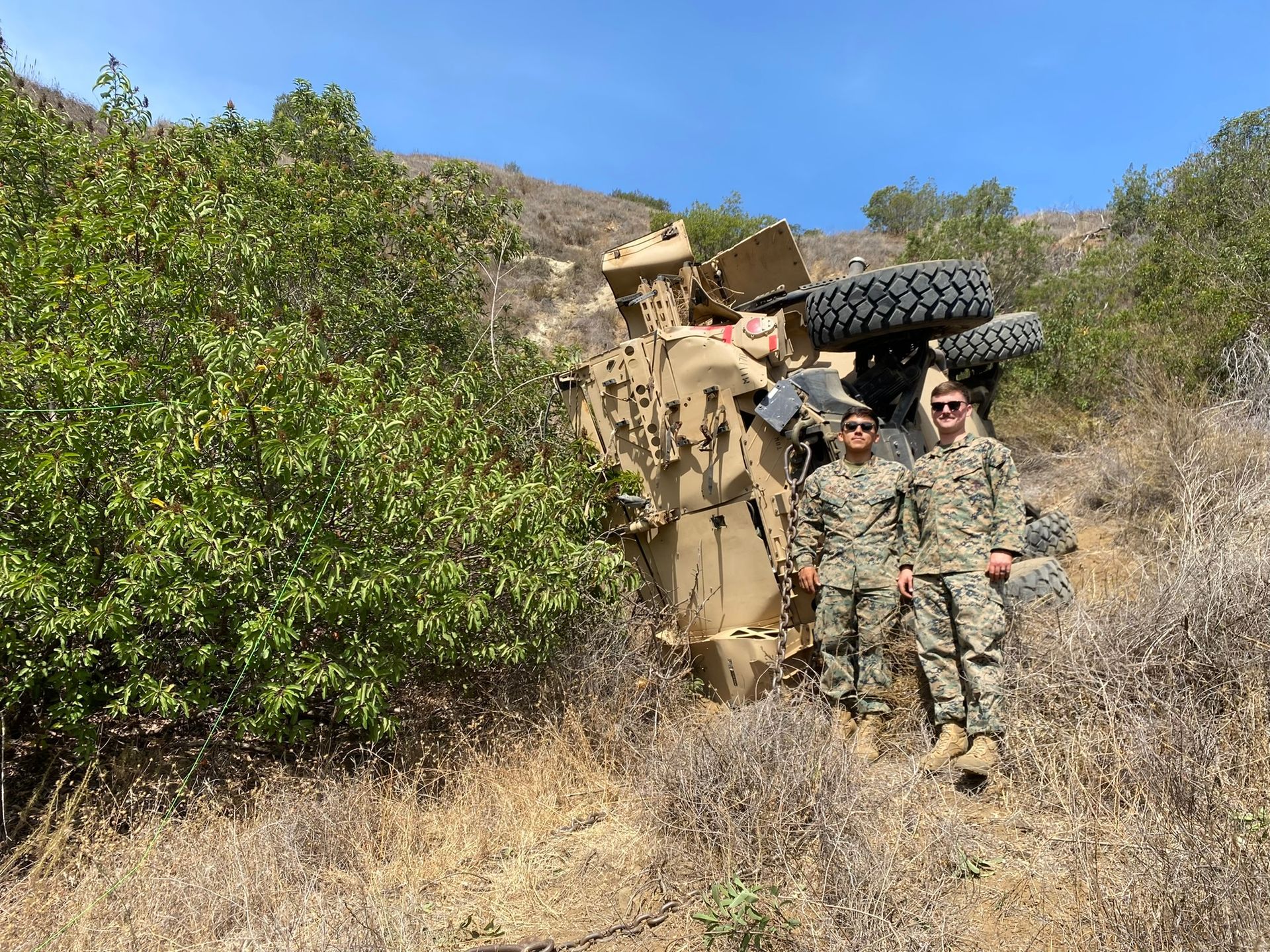 Two service members in camouflage uniforms stand smiling in front of an overturned military vehicle on a hillside.