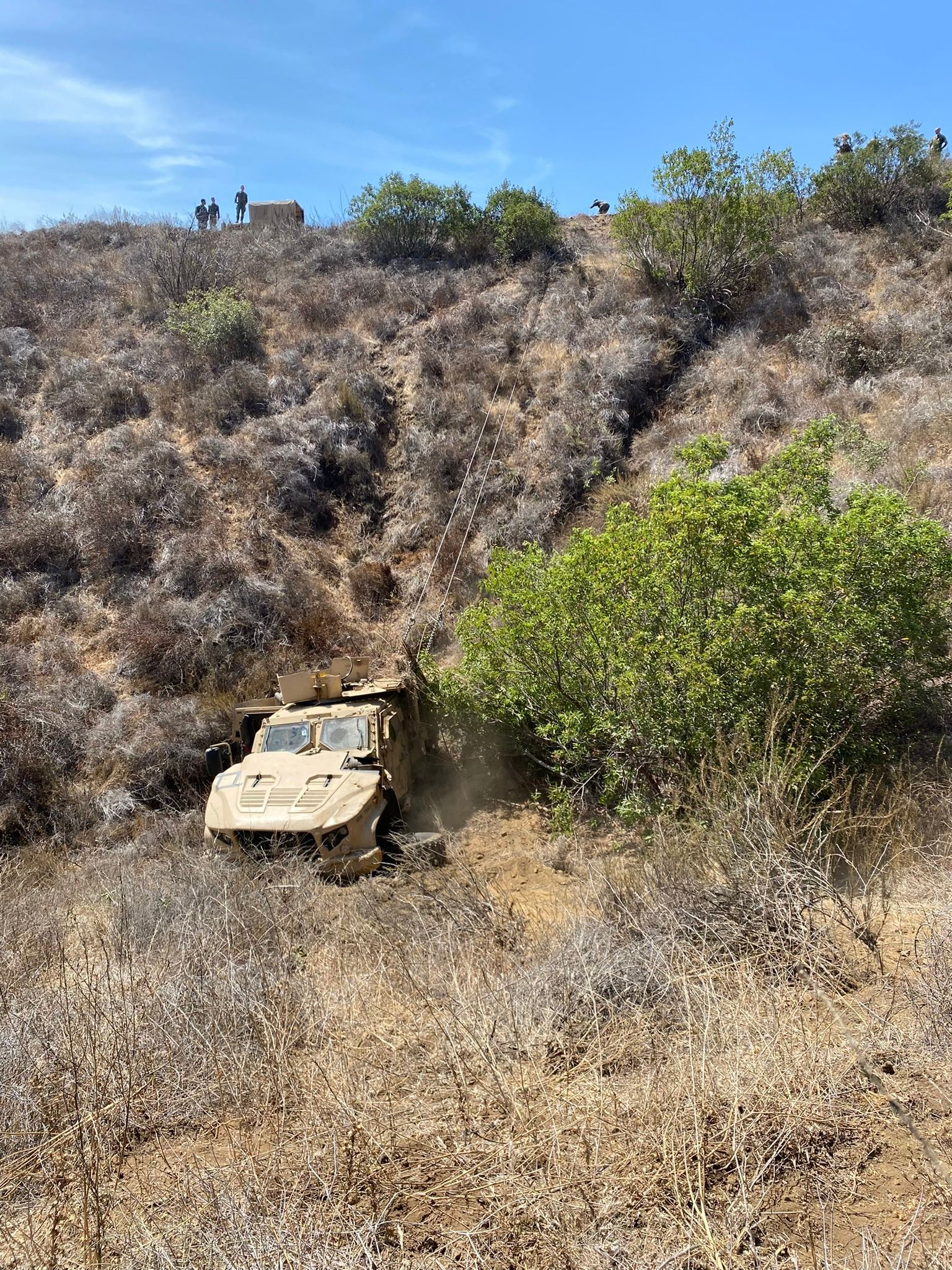 A tan military vehicle navigates a steep, dry, brush-covered hillside under a clear blue sky.