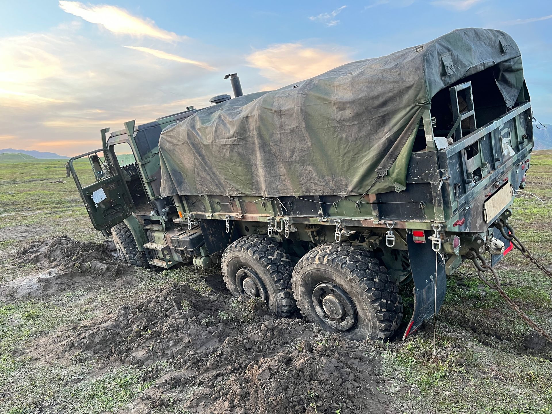 A green military truck is stuck in thick, dark mud in an open, grassy field under a cloudy sunset sky.