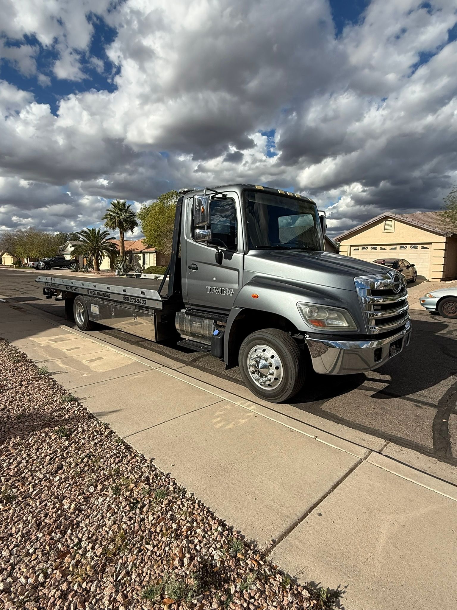 A silver Hino rollback tow truck parked on a residential street under a cloudy sky.