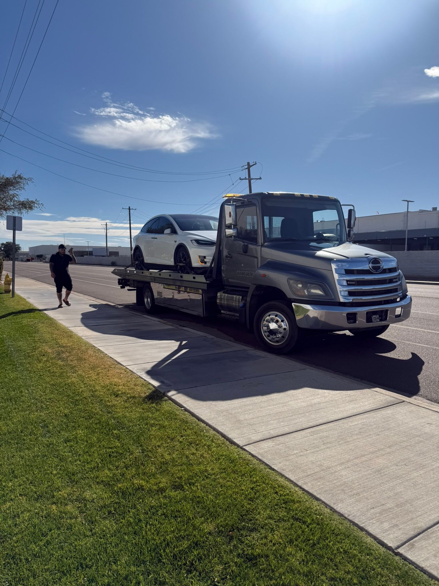 A white car is loaded onto a flatbed tow truck parked on a sunny street next to a sidewalk and grassy verge.