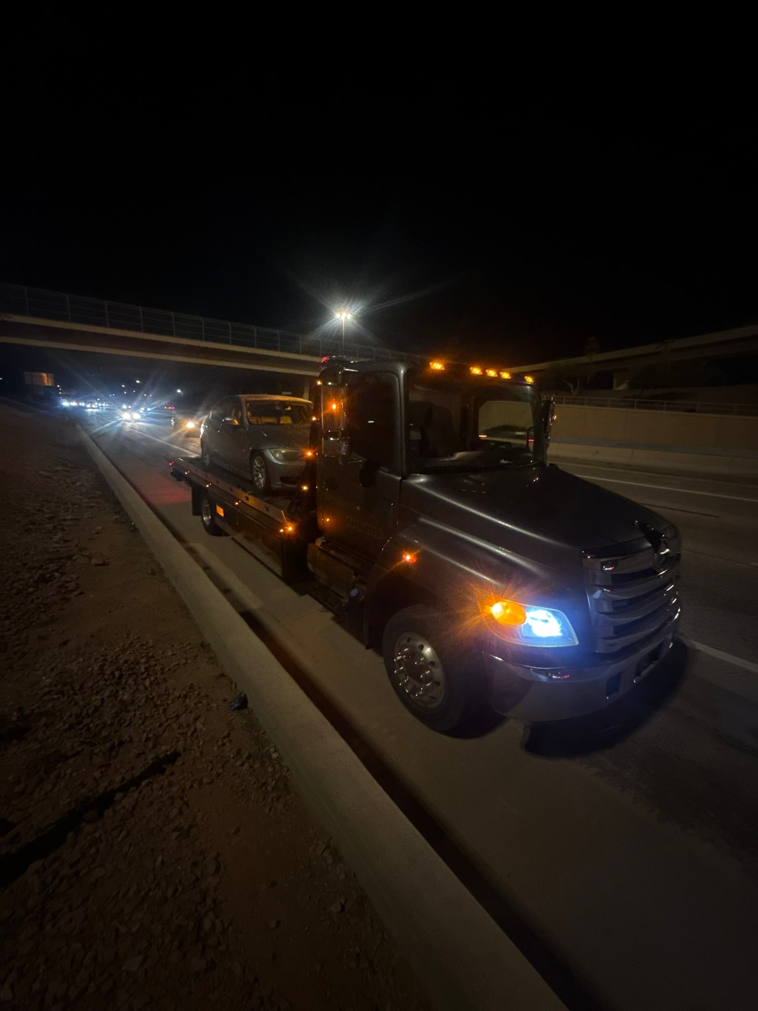 A flatbed tow truck with its headlights and orange marker lights on, carrying a car at night on a highway.