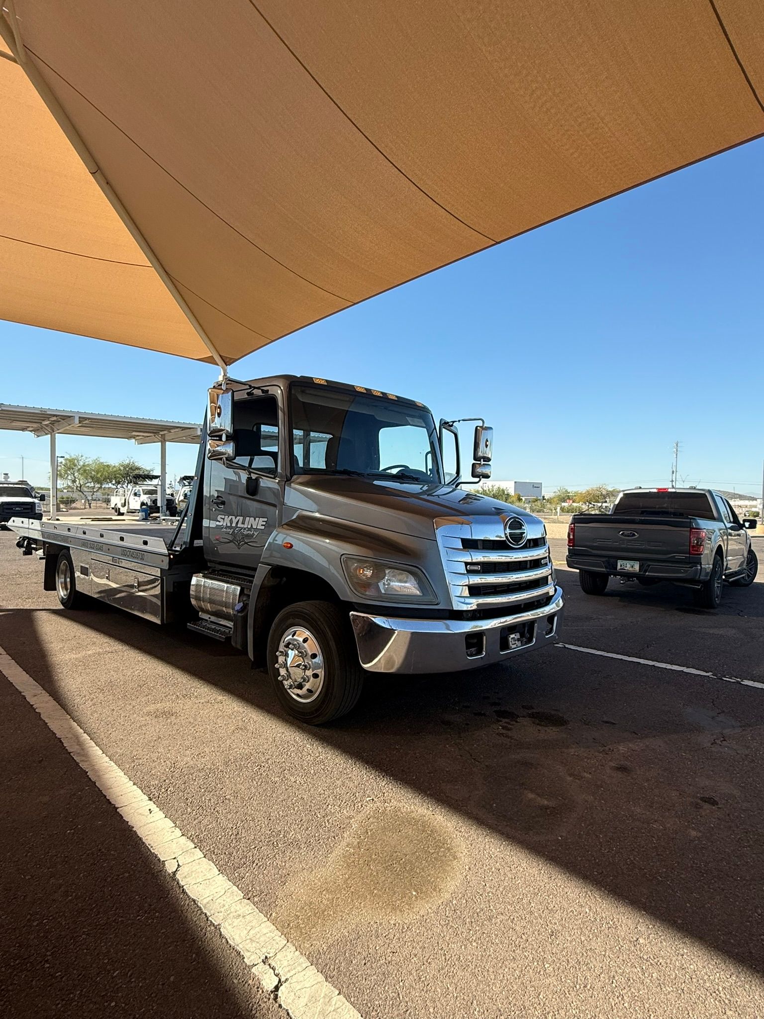 A silver flatbed tow truck parked under a shaded structure in a sunny paved parking lot, with a pickup truck nearby.