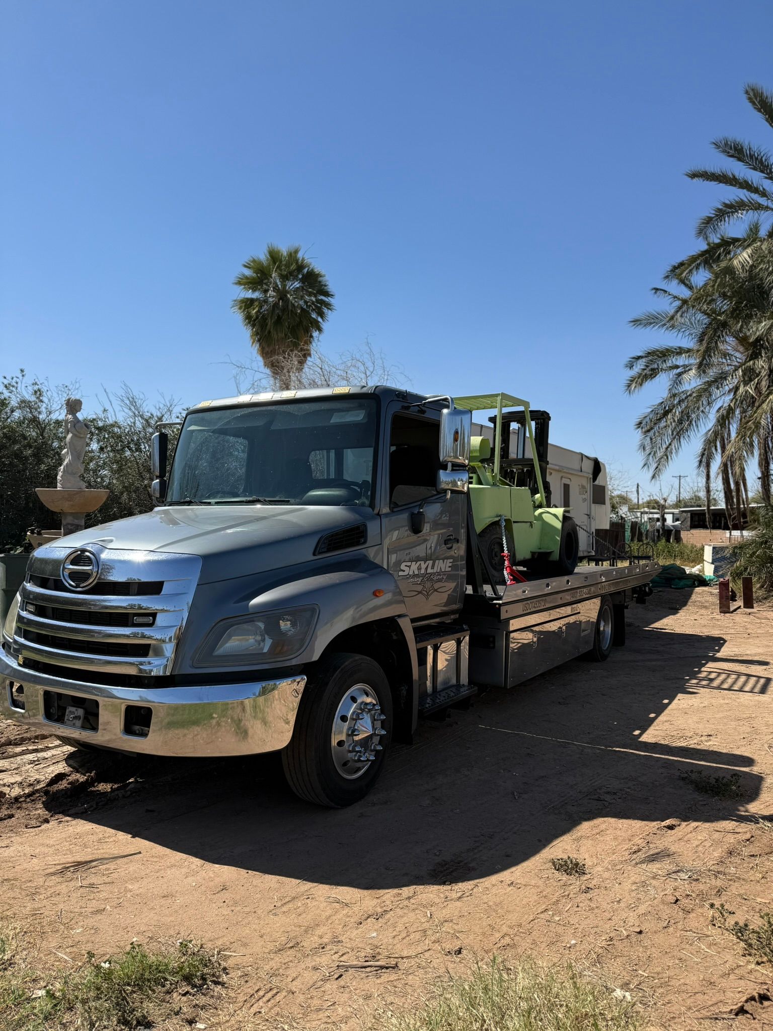 A grey flatbed tow truck carrying a light green forklift, parked on a dirt lot with palm trees in the background.