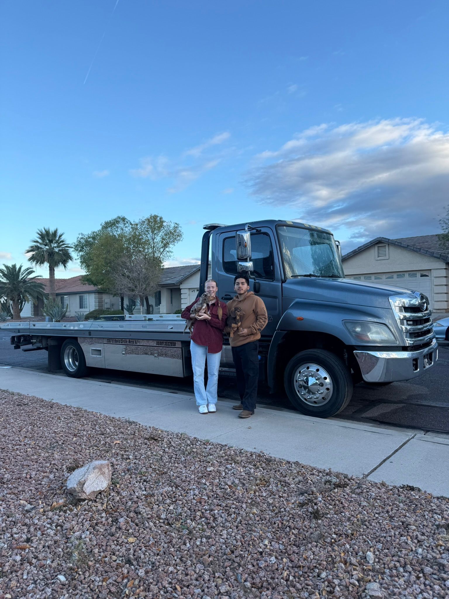 Two people stand smiling next to a large, grey flatbed tow truck parked on a suburban street under a blue sky.