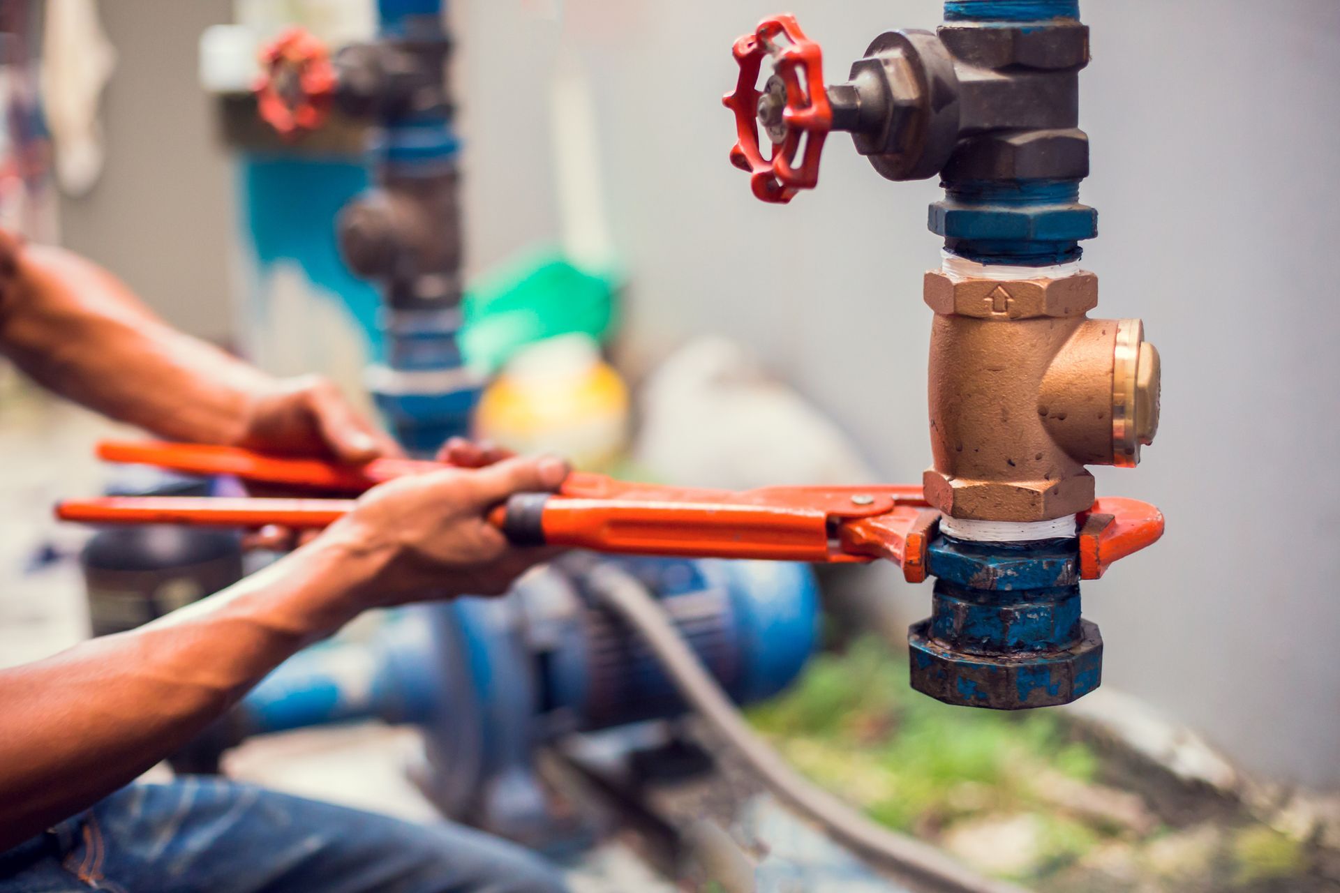A plumber uses an orange wrench to tighten a brass pipe connection, part of an outdoor water system.