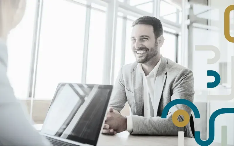 A man is shaking hands with another man while sitting at a table with a laptop.