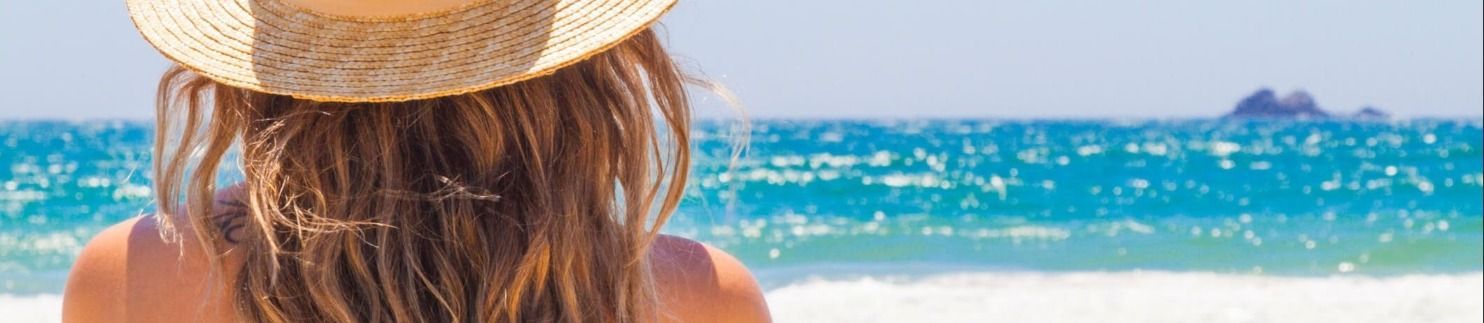 A woman wearing a hat is sitting on the beach looking at the ocean.