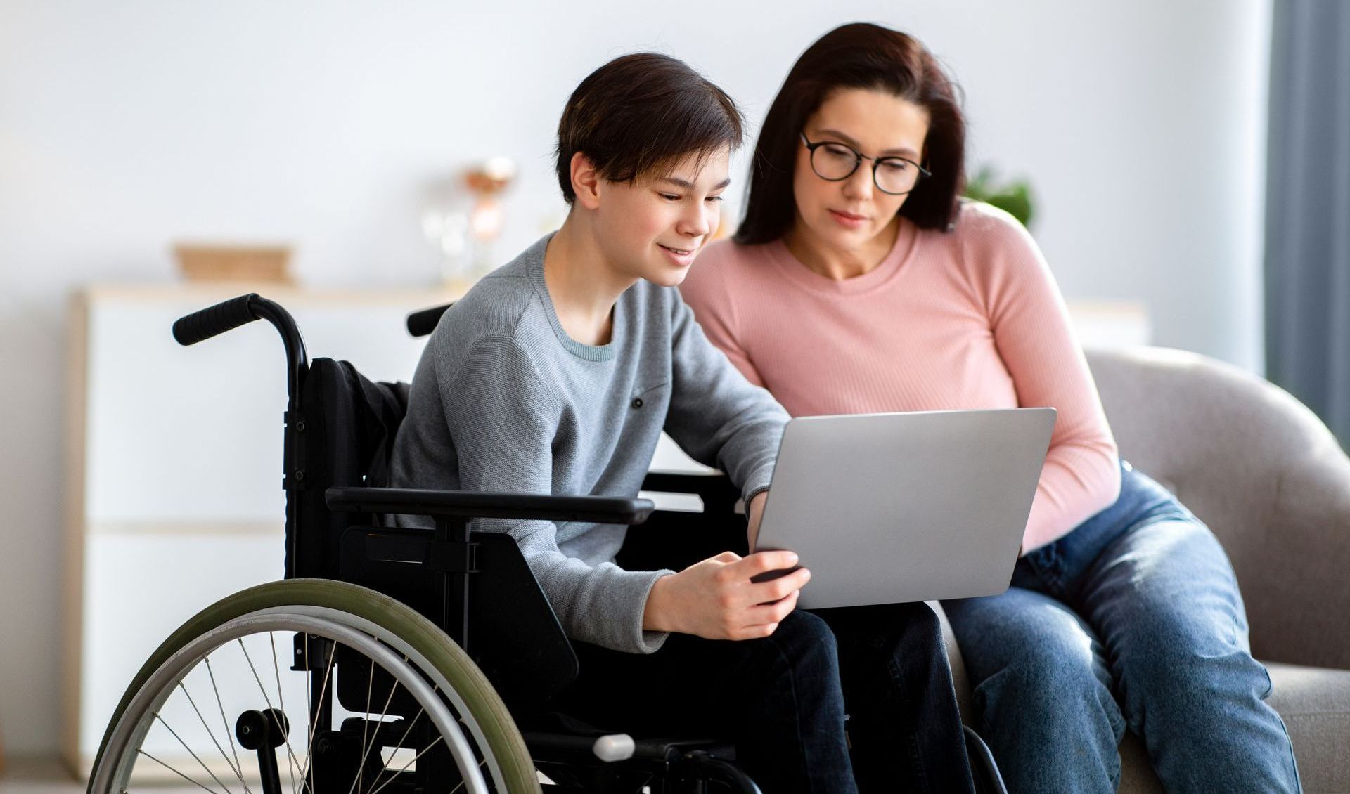 A woman is sitting next to a boy in a wheelchair using a laptop computer.