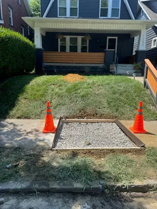 Sidewalk repair in progress: gravel-filled square with orange safety cones, front yard with grass and house 