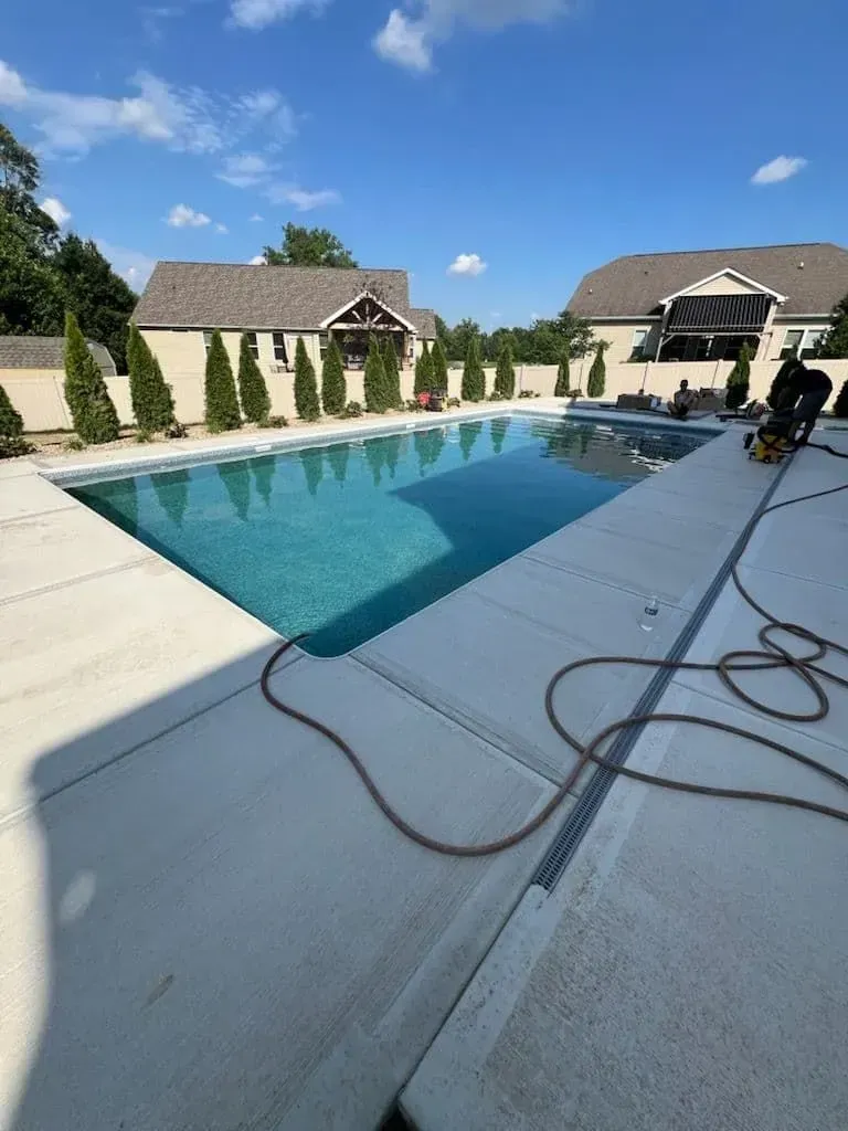 Rectangular swimming pool in a backyard with two houses and trees under a blue sky.