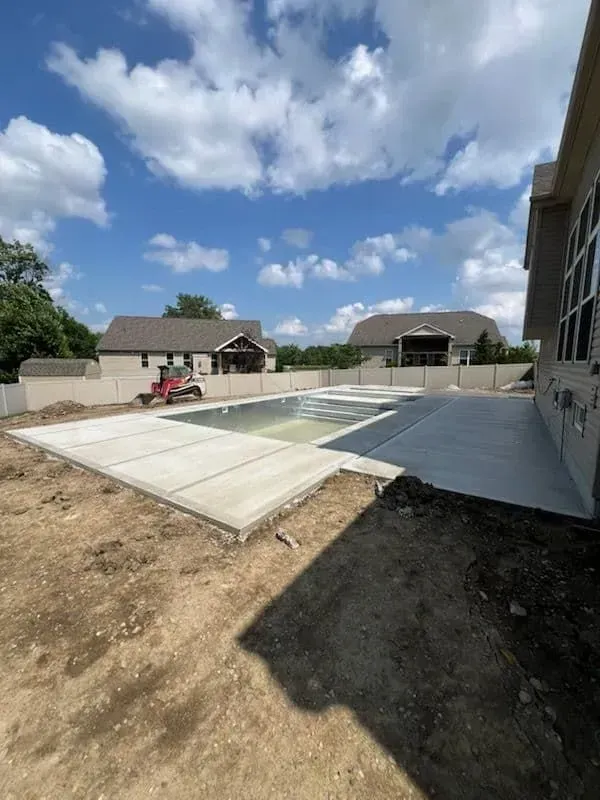 Concrete pool under construction with surrounding patio and two houses in the background. Blue sky with clouds.