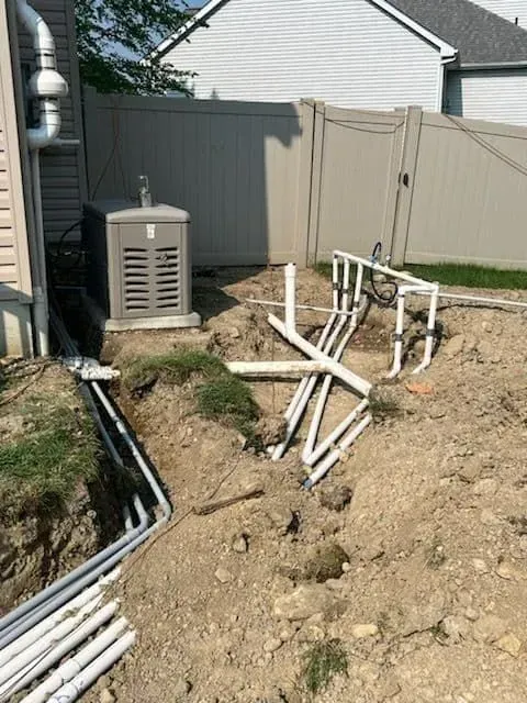 Backyard with buried PVC pipes, generator, and trench. Beige fence and house in the background.