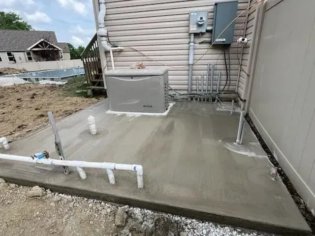Newly poured concrete pad with a generator, pipes, and electrical box by a beige building.