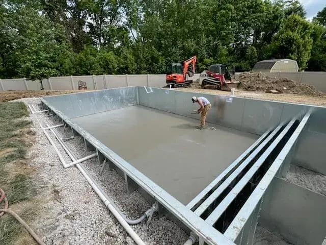 Man smoothing concrete in a rectangular pool under construction. Two excavators are in the background.