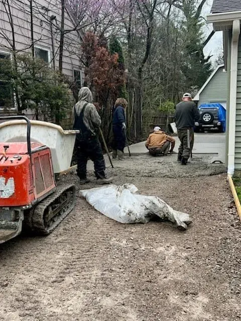 Construction workers paving a driveway with an excavator nearby and a car in an open garage.